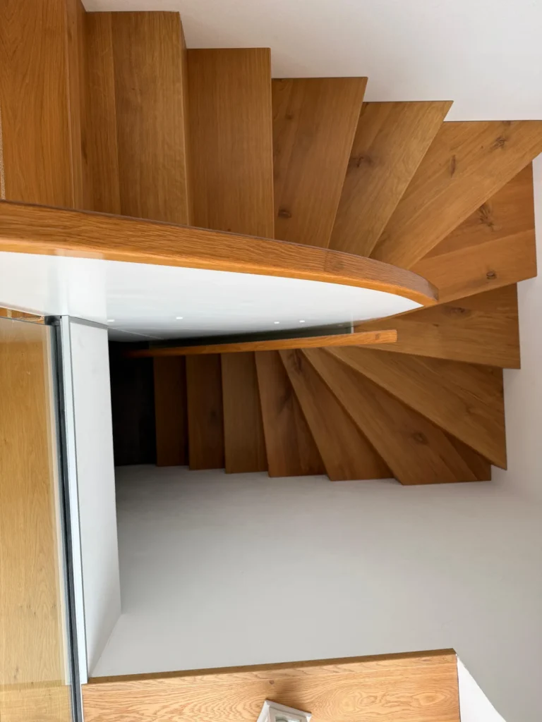 Spindeltreppe aus Holz, von oben gesehen, mit klaren Linien und hellbraunen Holzstufen, die teilweise von einer weißen Wand und einem Glasgeländer begrenzt werden.