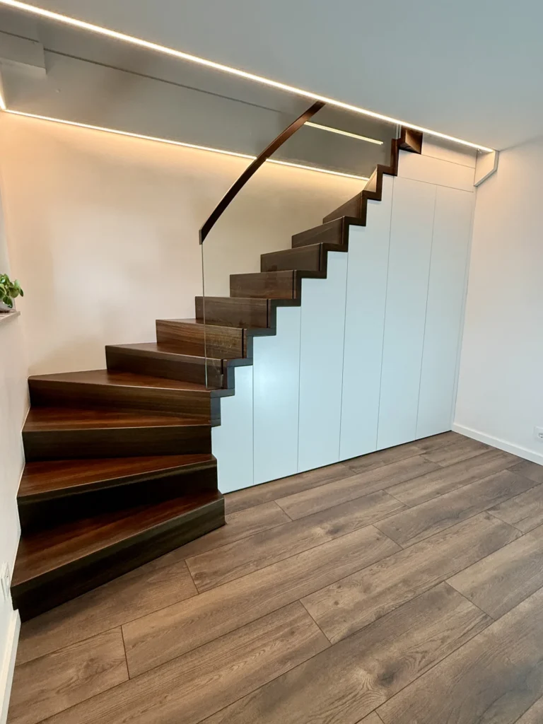 Eine moderne Treppe mit dunklen Holzstufen und einem schlichten Glasgeländer vor einer weißen Wand mit eingebauten Schränken darunter. Der Raum ist mit Holzfußböden und minimalistischen Designelementen ausgestattet.