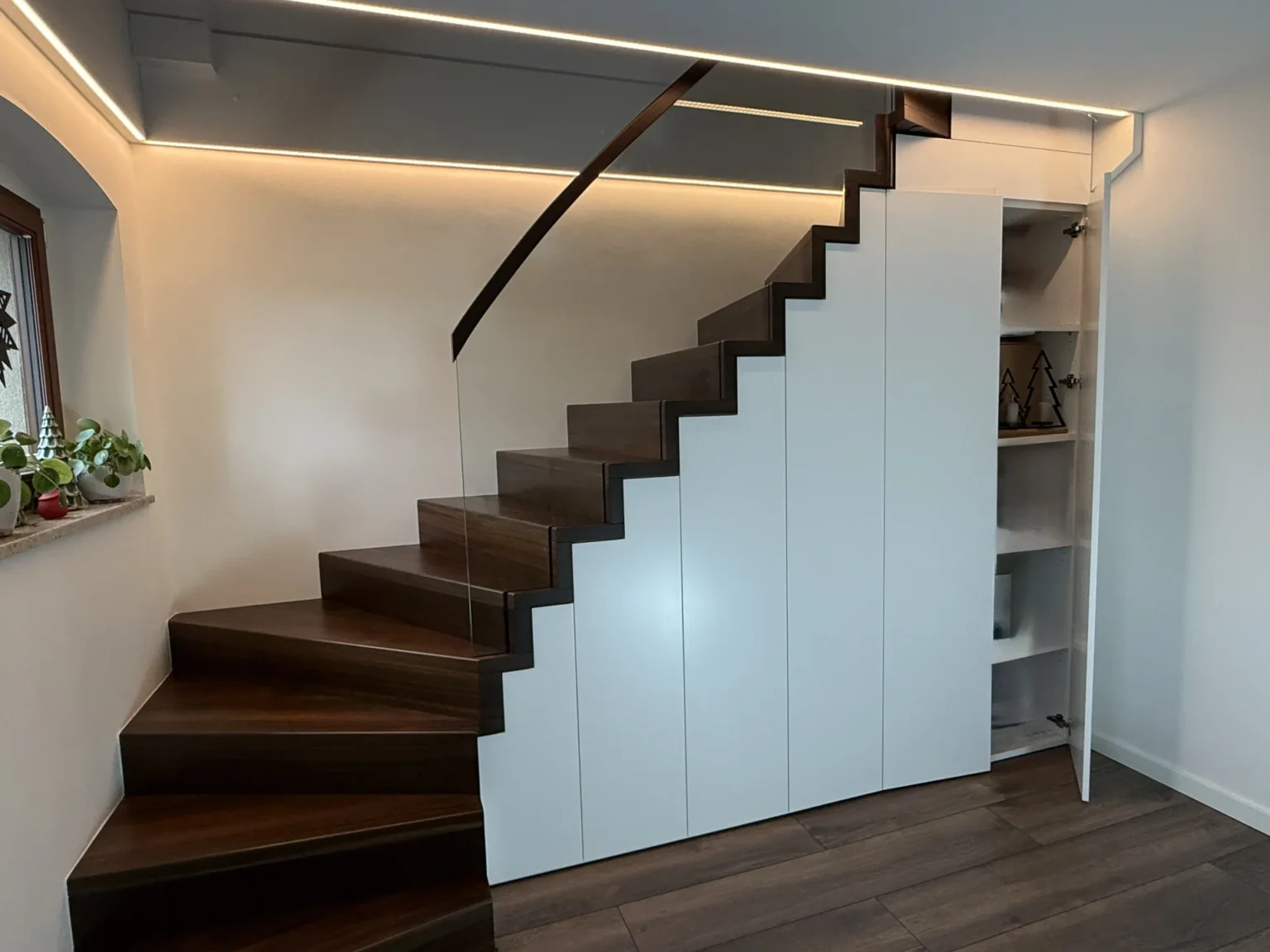 Moderne Holztreppe mit dunklen Stufen und darunter eingebauten weißen Schranktüren; eine Tür ist offen und gibt den Blick auf Regale mit Dekoration frei. An der Decke und am Treppengeländer verlaufen LED-Lichtbänder.