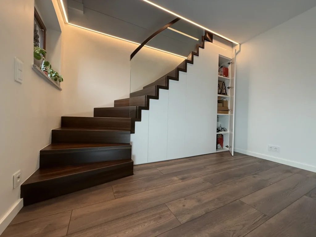 Eine moderne Holztreppe mit dunklen Stufen und weißem Sockel schlängelt sich neben einer Wand nach oben. Unter der Treppe sind Regale mit Büchern und Dekoration aufgebaut. Der Raum hat Holzböden und eine sanfte LED-Beleuchtung.