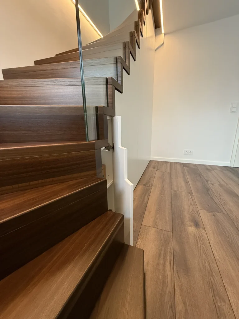 Eine moderne Treppe mit dunklen Holzstufen, einem Glasgeländer und einer weißen Wand daneben, in einem Raum mit passendem Holzboden und minimalistischen weißen Wänden.