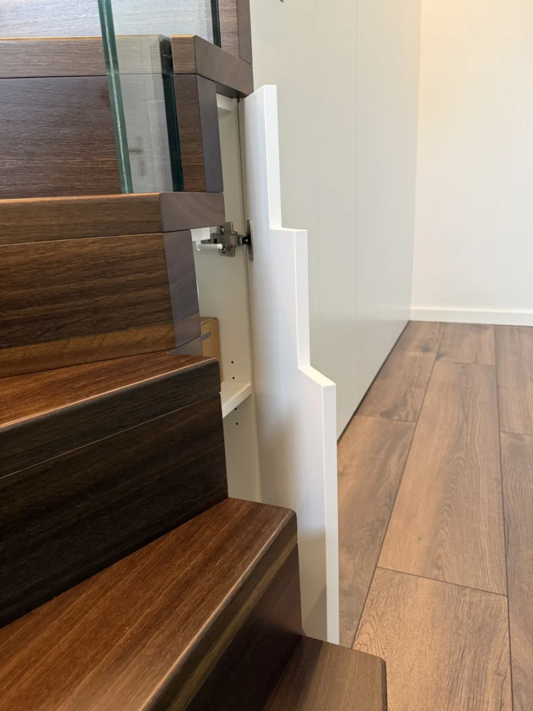 Ein versteckter Schrank ist in die Seite einer Treppe mit dunklen Holzstufen eingebaut, mit einer weißen Tür, die zum Treppenprofil passt und sich öffnen lässt, um Regale im Inneren freizugeben.