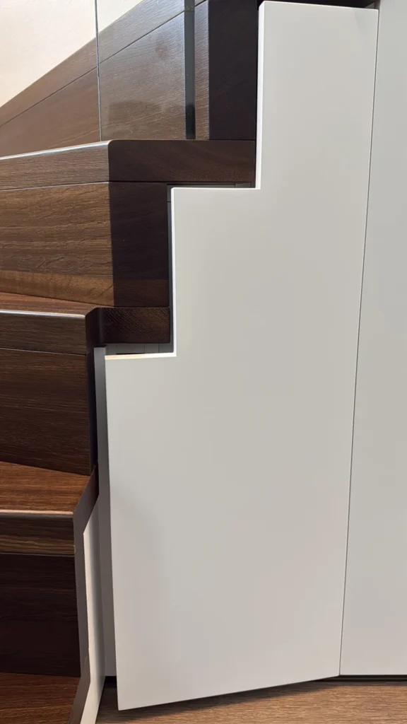 Nahaufnahme einer modernen Holztreppe mit einer weißen Schranktür, die eine abgestufte Kante hat, die an die Kontur der darüber liegenden Treppe angepasst ist.