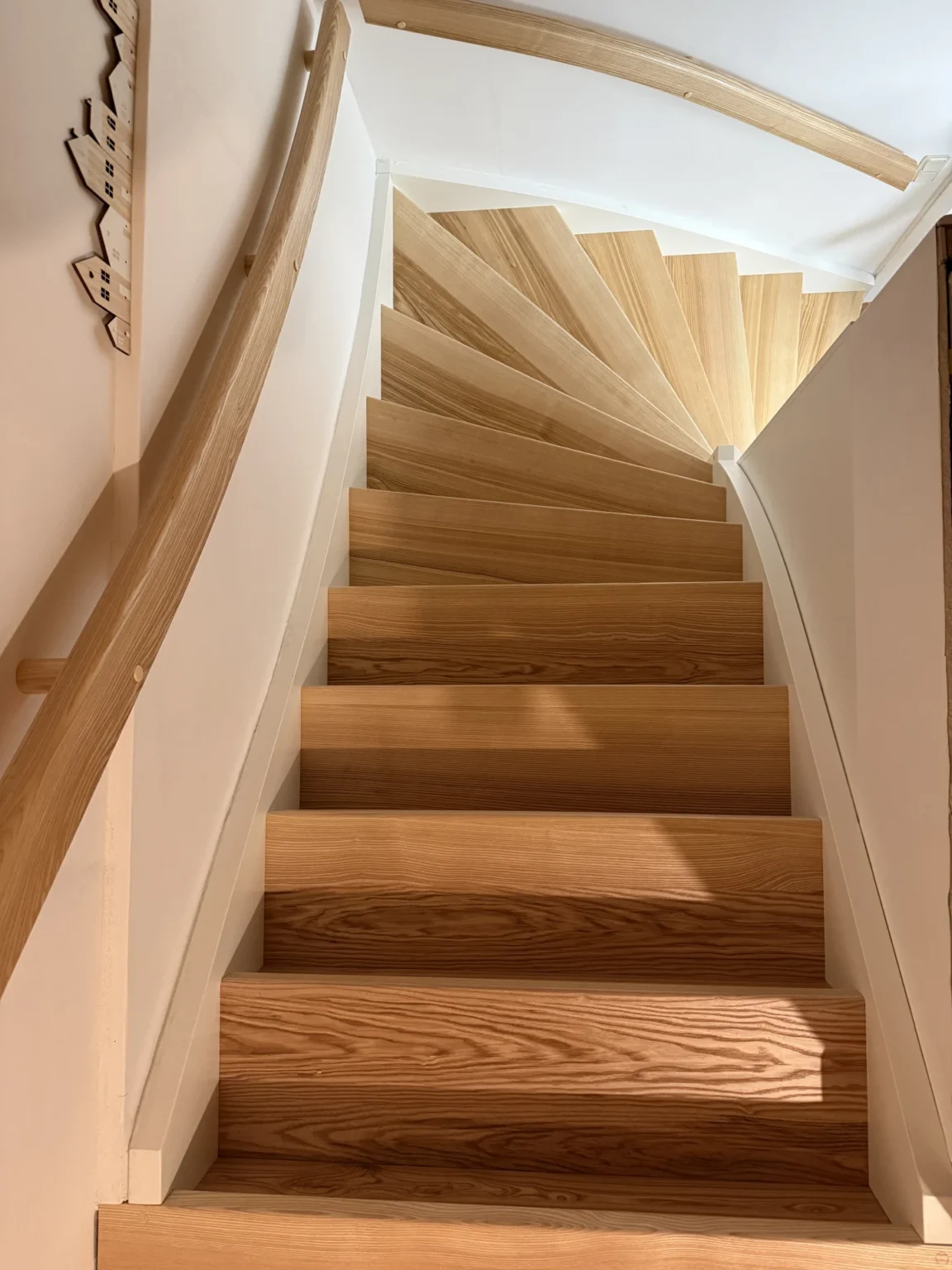 Eine Holztreppe mit hellen Stufen und einem geschwungenen Handlauf führt spiralförmig nach oben und wird von weißen Wänden in einem hellen Innenraum begrenzt.