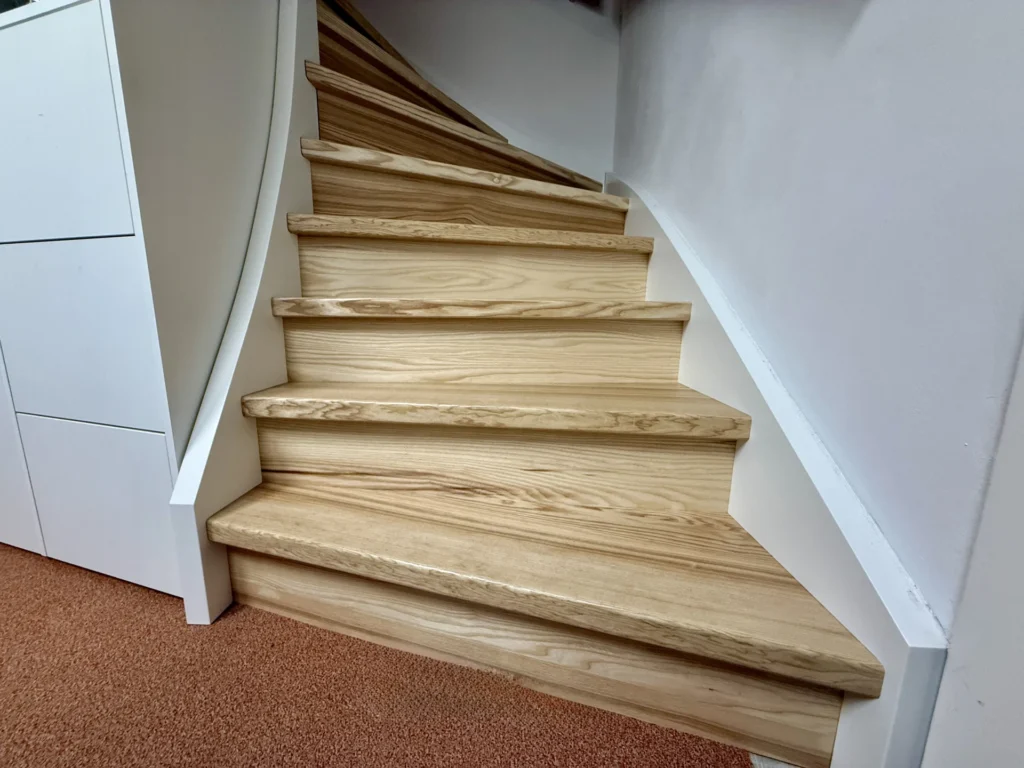 Eine helle, leicht geschwungene Holztreppe, die von weißen Wänden und einem braunen Teppichboden am Fußende begrenzt wird.