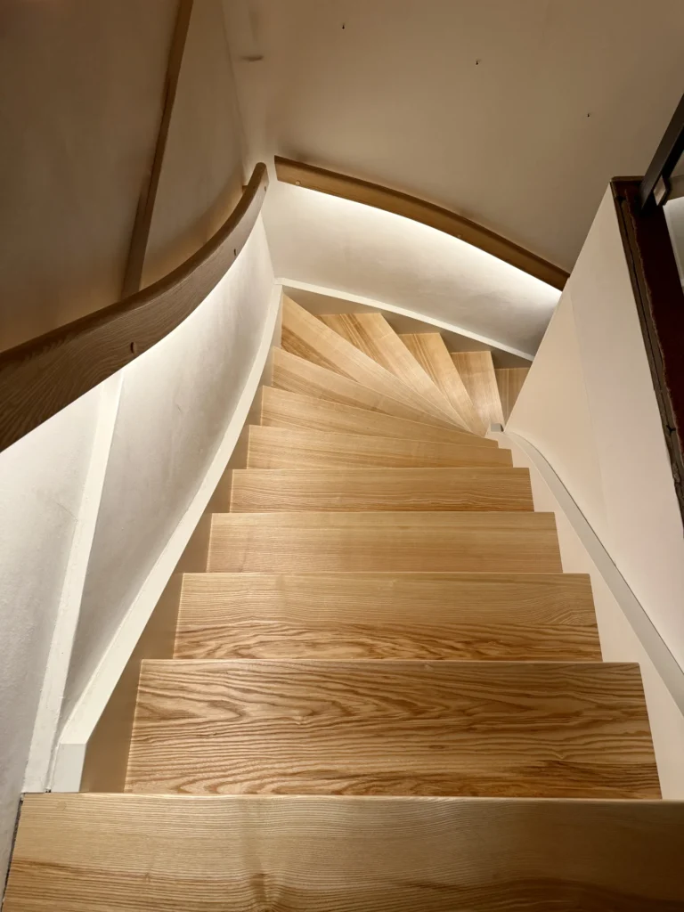 Eine Wendeltreppe aus hellem Holz schlängelt sich nach unten, begrenzt von weißen Wänden und einem schlichten Holzgeländer, was ein modernes und elegantes Design ergibt.