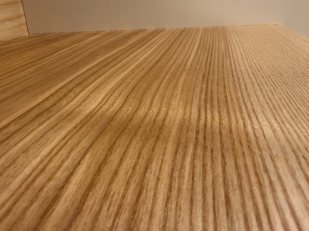 Nahaufnahme einer Holzoberfläche mit ausgeprägter, wellenförmiger Maserung, die diagonal über das Bild verläuft. Das Holz erscheint glatt und hell in der Farbe, mit einem einfachen, minimalistischen Hintergrund.