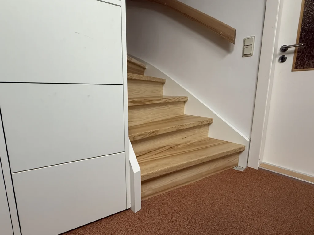 Eine kleine, geschwungene Treppe mit hellen Holzstufen und einem Holzhandlauf führt von einem Raum mit braunem Teppich und weißen Wänden nach oben. Neben der Treppe stehen weiße Einbauschränke.