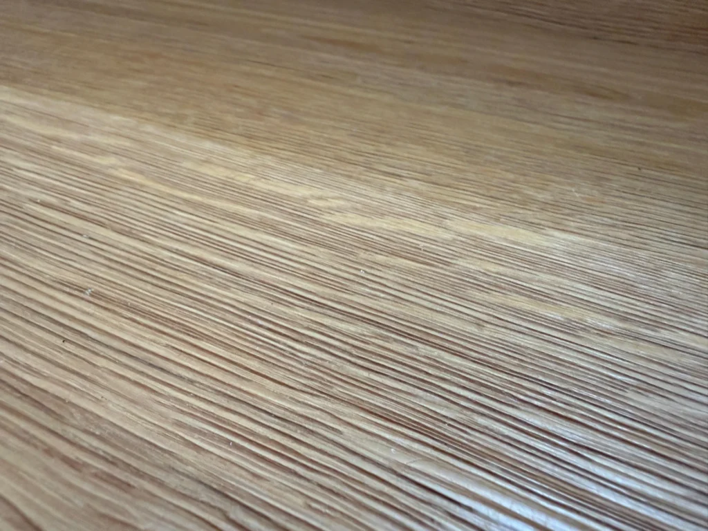Nahaufnahme einer Holzoberfläche mit sichtbarer Maserung und Textur, die parallele Linien und eine natürliche hellbraune Färbung aufweist.