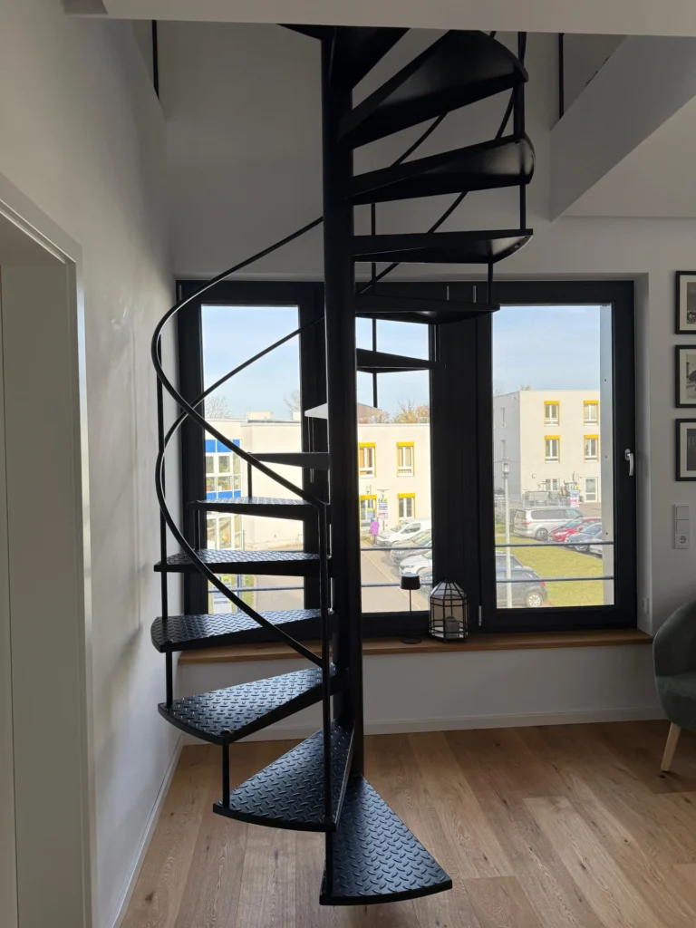 Eine schwarze Wendeltreppe aus Metall steht in einem modernen Raum mit hellem Holzboden und großen Fenstern, durch die man Gebäude und geparkte Autos von außen sehen kann.