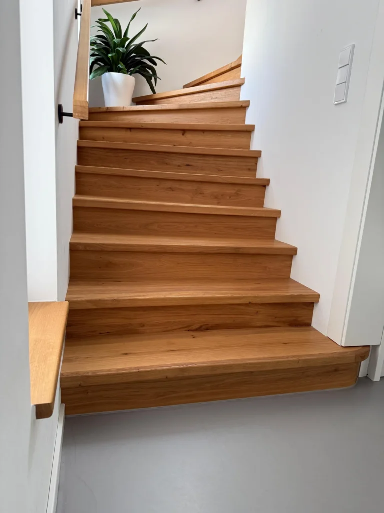 Eine moderne Holztreppe mit hellbraunen Stufen führt zickzackförmig nach oben. Eine weiße Wand und ein schwarzer Handlauf säumen die Treppe, und eine grüne Topfpflanze steht auf dem oberen Treppenabsatz.