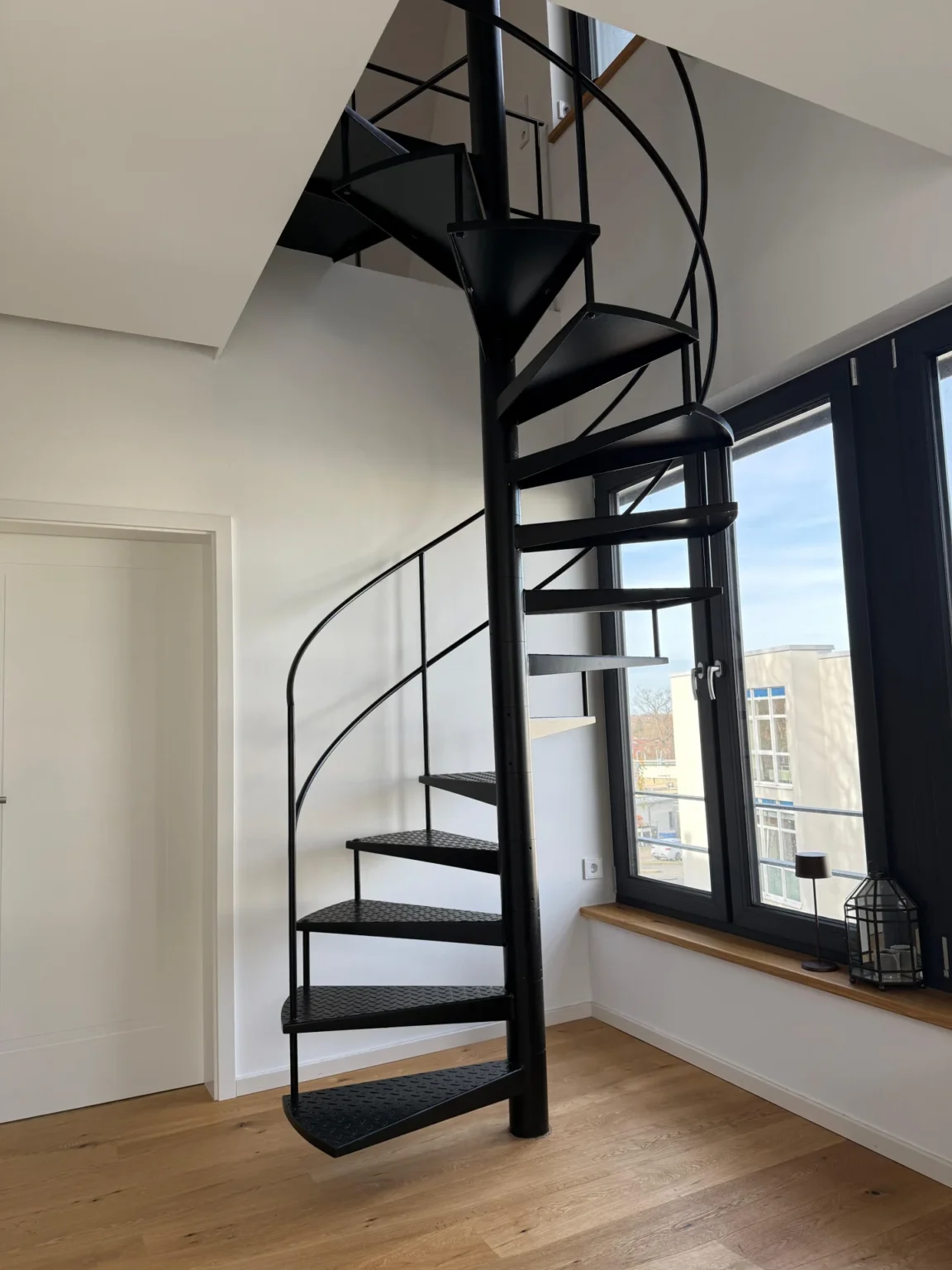 Eine schwarze Wendeltreppe aus Metall steht in einem hellen Raum mit Holzböden, weißen Wänden und großen Fenstern, die natürliches Licht hereinlassen.