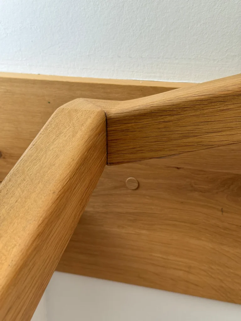 Nahaufnahme eines hölzernen Handlaufs mit einer präzisen, abgewinkelten Verbindung, montiert auf einer passenden Holzplatte, die an einer weißen Wand befestigt ist. Das Holz hat eine natürliche Oberfläche, die seine Maserung und Textur zur Geltung bringt.