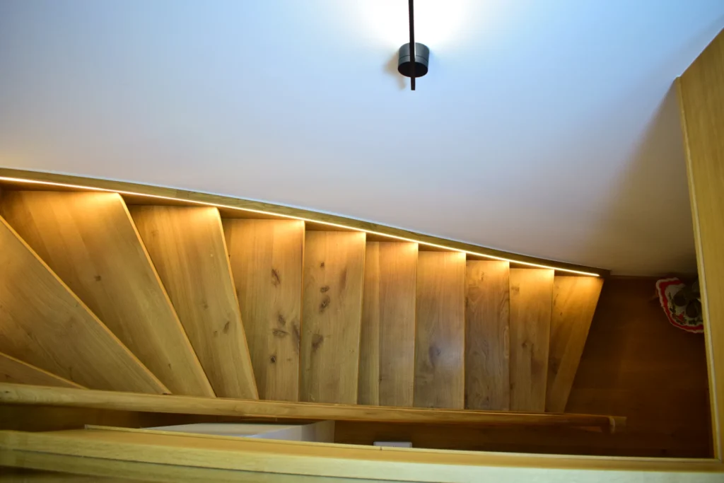 Eine hölzerne Wendeltreppe mit eingebauten LED-Lichtbändern an der Wand, von oben gesehen unter einer Deckenleuchte.