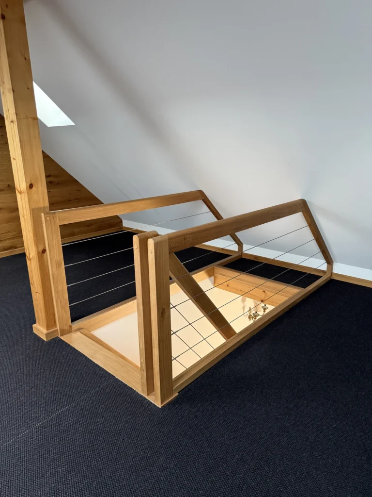 Ein hölzernes Treppengeländer mit Metallseilen umschließt eine Öffnung in einem mit Teppich ausgelegten Dachboden, die zu einer Treppe darunter führt. Die Wände sind weiß, und natürliches Licht fällt durch ein Oberlicht ein.