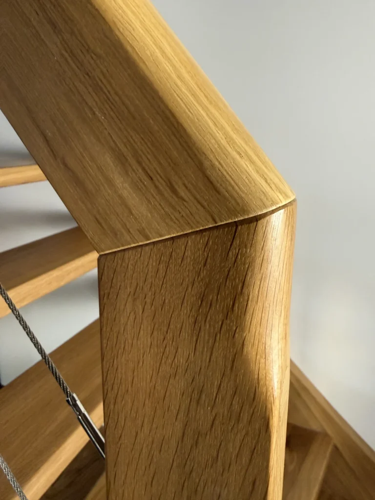 Nahaufnahme eines hölzernen Treppengeländers mit einer genau abgewinkelten Verbindung. Das Holz ist glatt und poliert, und ein Metallkabel verläuft parallel zum Geländer unter ihm. Der Hintergrund ist eine schlichte, helle Wand.