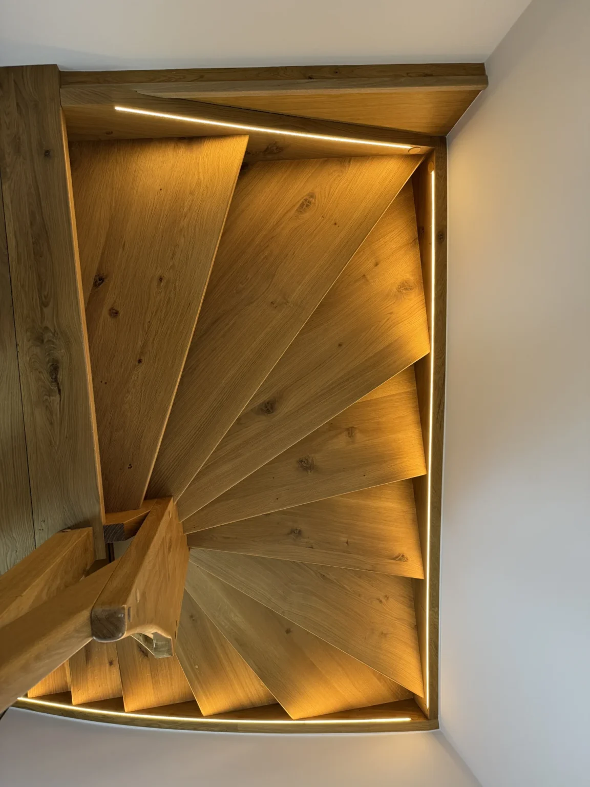 Draufsicht auf eine moderne Holzspindeltreppe mit integrierter warmer LED-Beleuchtung, die jede Stufe hervorhebt und ein geometrisches Muster erzeugt.