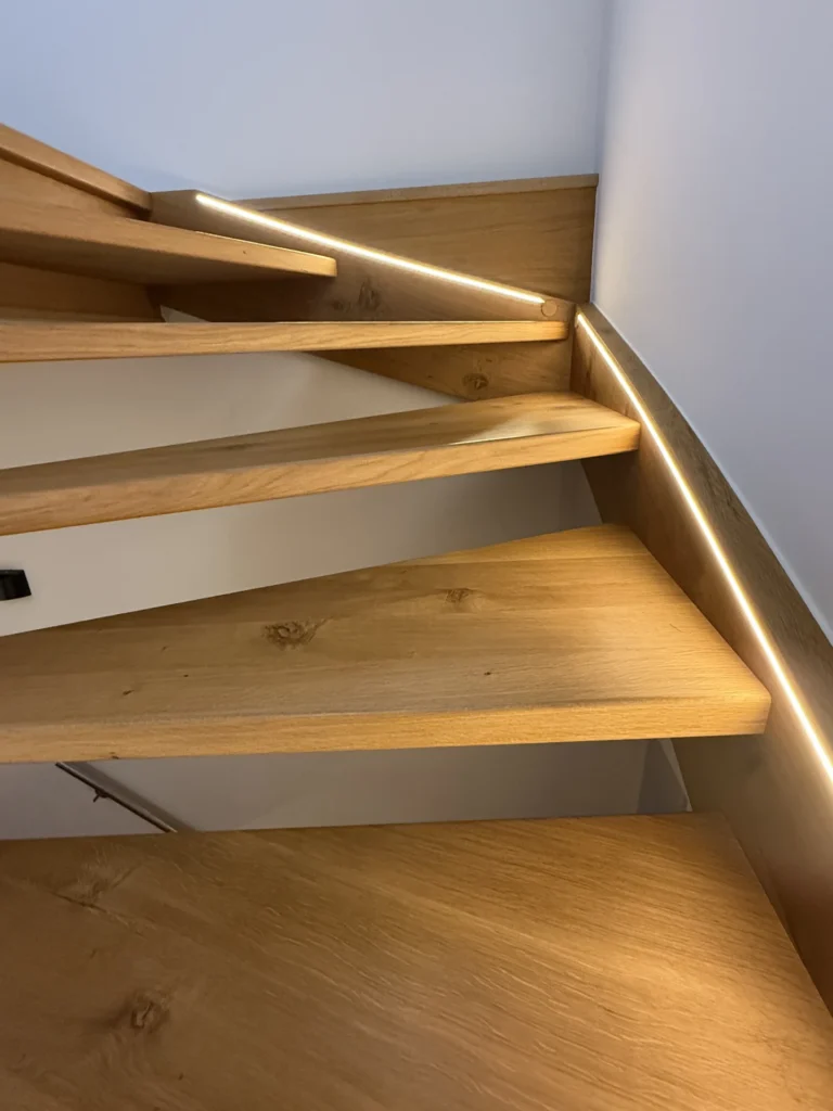 Moderne Holztreppe mit schwimmenden Stufen und eingebauten LED-Lichtbändern entlang der Wand, die ein warmes und modernes Aussehen schaffen. Die Stufen sind eckig und aus hellem Holz gefertigt.
