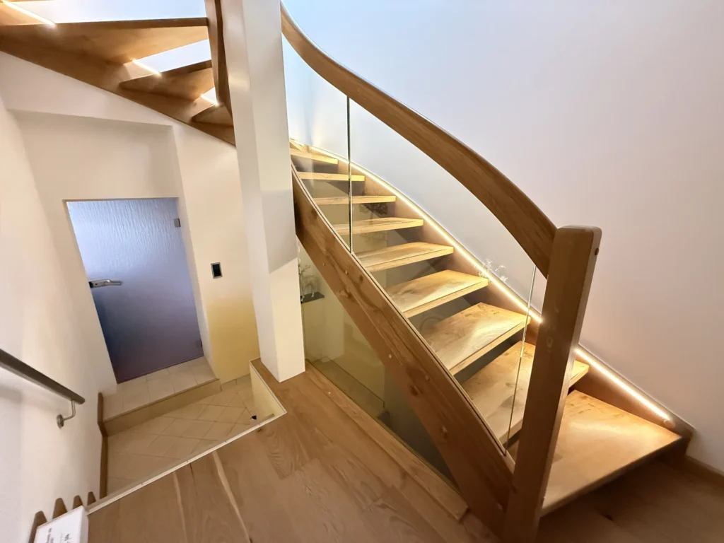 Eine moderne Holztreppe mit Glaspaneelen und eingebauter LED-Beleuchtung schlängelt sich in einem hellen Innenraum nach oben. Die Stufen und das Geländer sind aus Holz, und am unteren Ende der Treppe ist eine blaue Tür zu sehen.