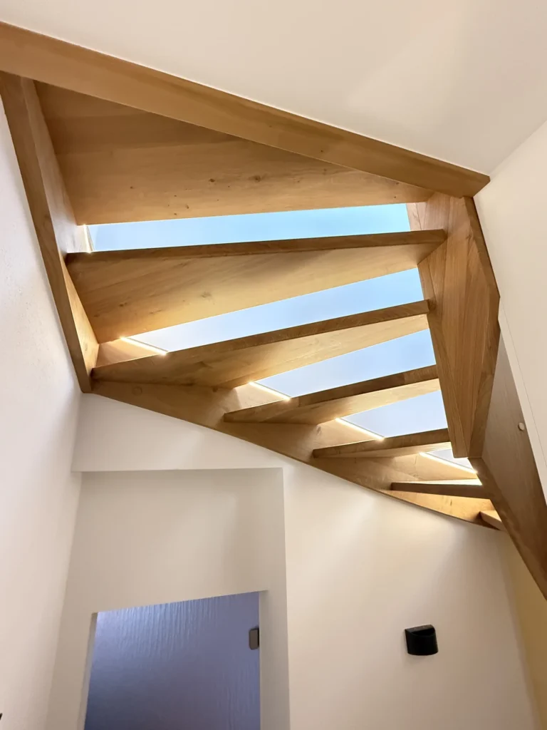 Ein Blick auf eine moderne Holztreppe mit kantigen Balken und einem Oberlicht darüber, das natürliches Licht in einen weißwandigen Innenraum lässt.