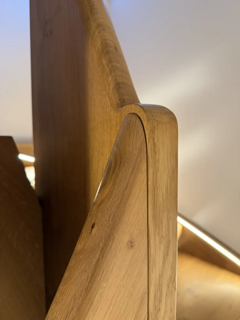 Nahaufnahme eines Treppenhandlaufs aus Holz mit glatten, abgerundeten Kanten und natürlicher Holzmaserung, der sich durch moderne Handwerkskunst auszeichnet und durch sanfte Beleuchtung beleuchtet wird.