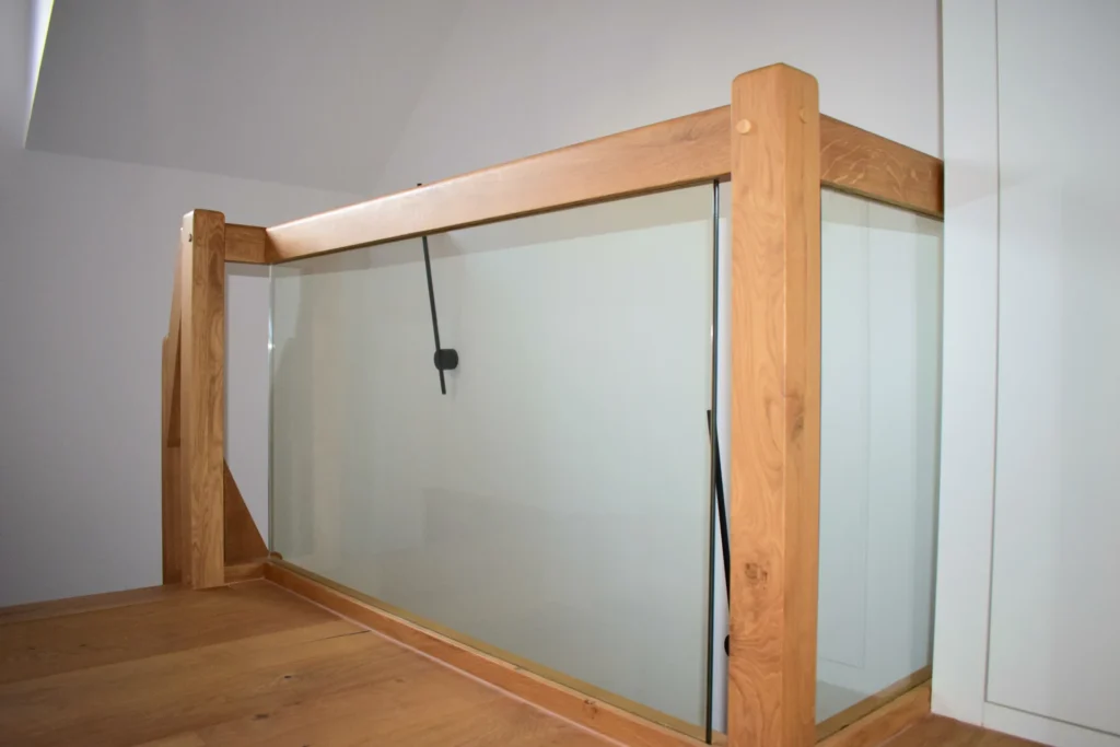 Abgebildet ist ein modernes Treppengeländer aus Klarglas mit Holzpfosten und Handläufen vor einer hellen Wand und einem Hartholzboden. An der Innenseite des Geländers hängt ein schwarzes Kabel oder eine Schnur.