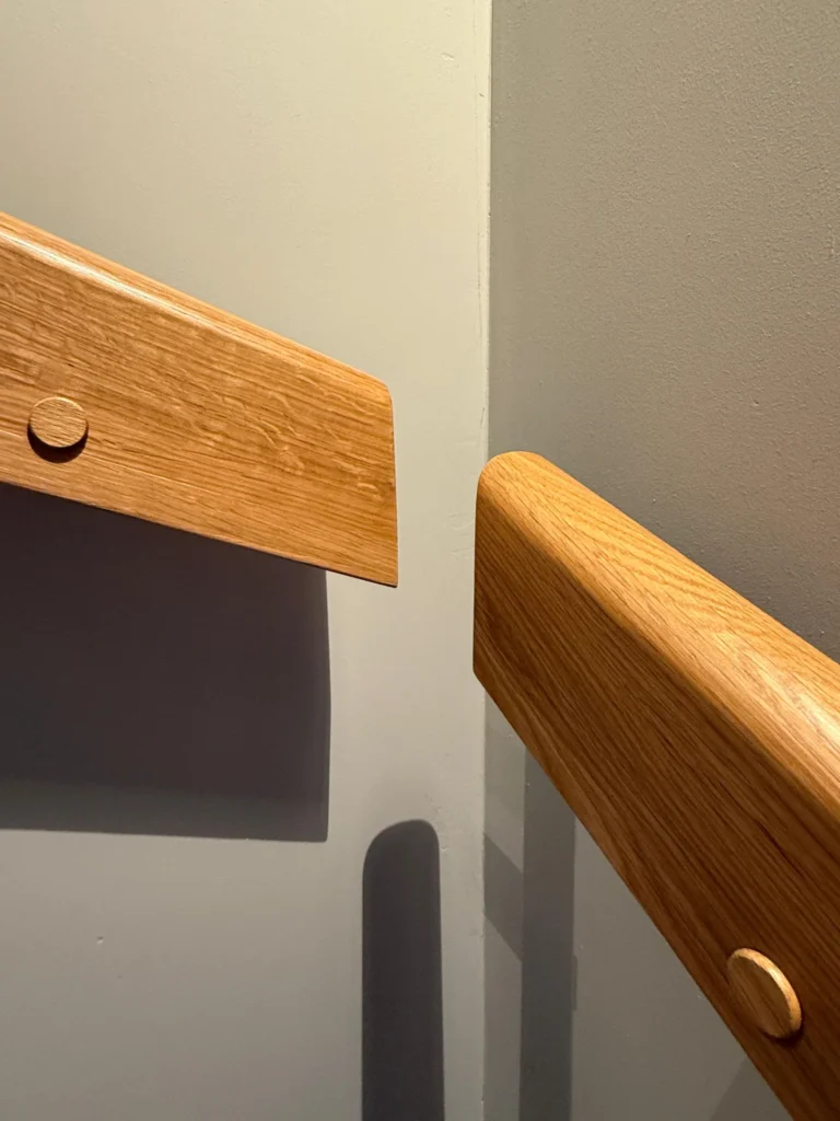 Zwei hölzerne Handläufe mit abgerundeten Enden sind an gegenüberliegenden Wänden in einer Ecke angebracht und berühren sich nur knapp. Die Wände sind hellgrau und bilden eine einfache, minimalistische Komposition.