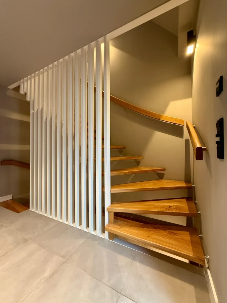 Moderne Holztreppe mit offenen Setzstufen und schlichter weißer vertikaler Lamellentrennwand, vor beigen Wänden und großen hellen Bodenfliesen, mit minimalistischer Beleuchtung an der Wand.