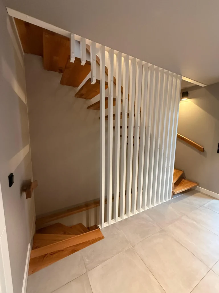 Eine moderne Treppe mit Holzstufen und einer vertikalen Trennwand aus weißen Lamellen vor hellen Wänden und einem gefliesten Boden in einem zeitgenössischen Innenraum.