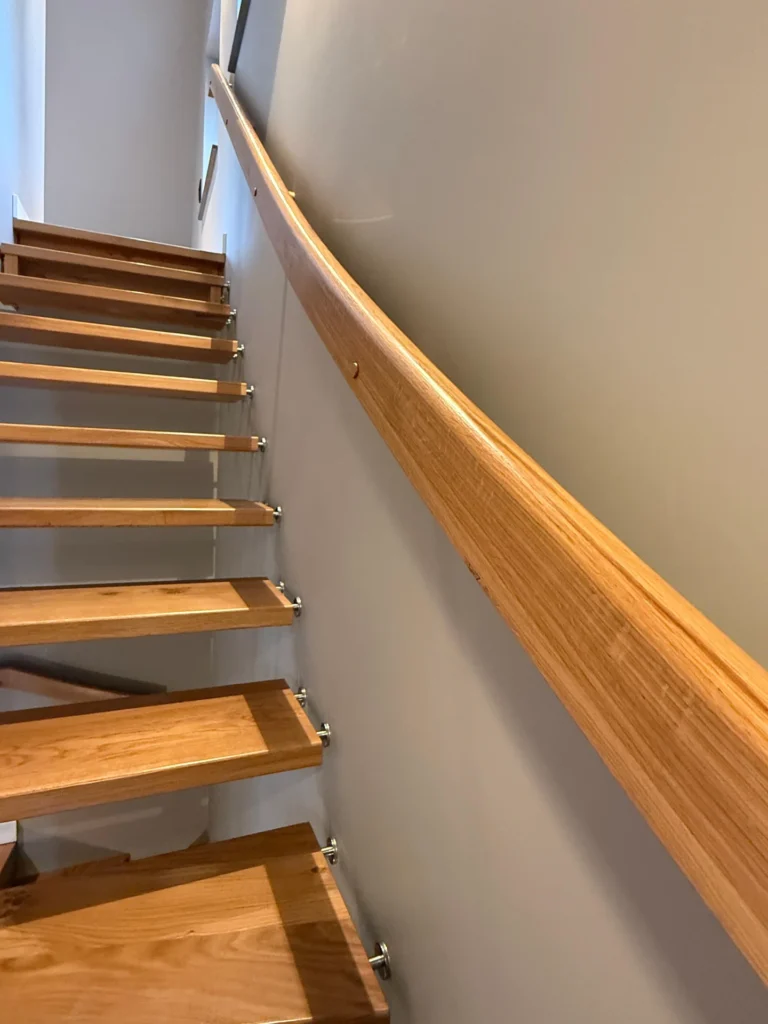 Eine moderne Holztreppe mit offenen Setzstufen und einem eleganten Holzhandlauf an einer hellen Wand, die in einem gut beleuchteten Innenraum nach oben führt.