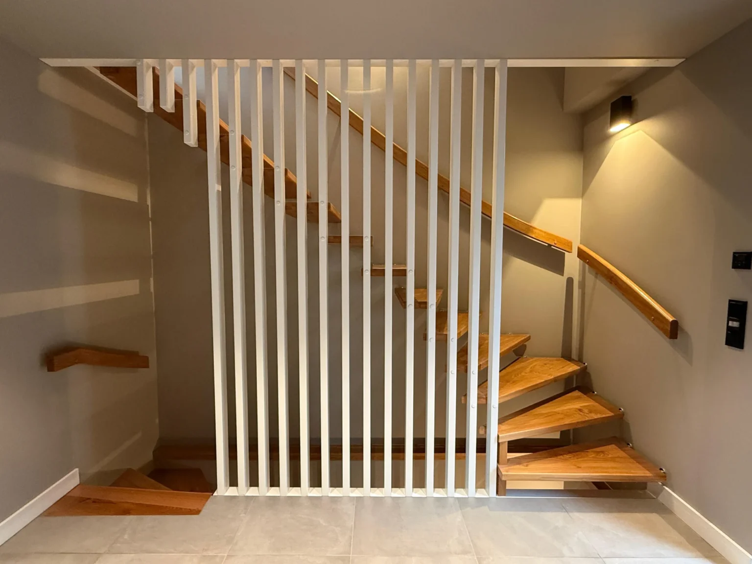 Moderne Holztreppe mit kantigen Stufen und schlanken Handläufen, die teilweise von vertikalen weißen Latten eingefasst sind, vor hellen Wänden und gefliestem Boden in einem minimalistischen Innenraum.