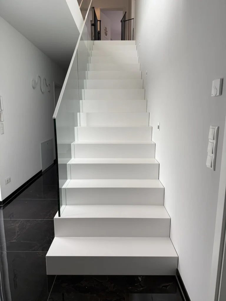 Moderne weiße Treppe mit glattem Glasgeländer in einem minimalistischen Interieur mit schwarzem Fliesenboden und weißen Wänden. Die Treppe führt in ein Obergeschoss mit einem Flur und einem Geländer am oberen Ende.