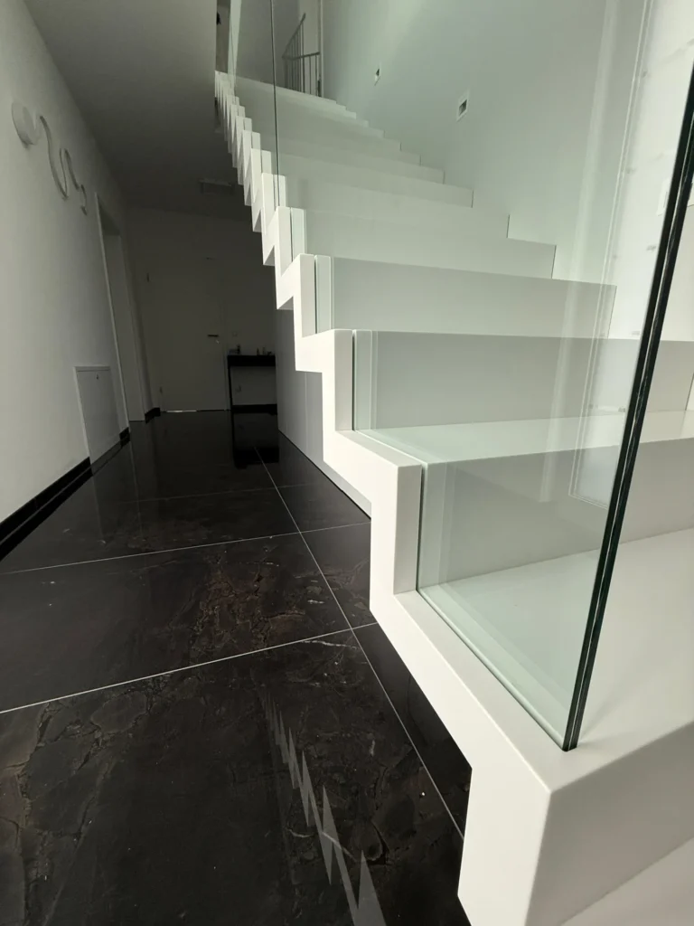 Ein modernes Interieur mit einer weißen, schwebenden Treppe mit Glasgeländer und dunklen, glänzenden Bodenfliesen. Der Raum ist minimalistisch mit klaren Linien und weißen Wänden.