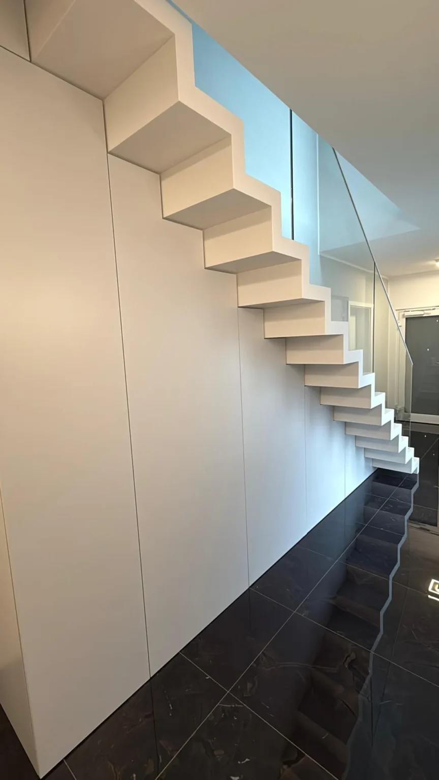Minimalistische, moderne weiße Treppe mit scharfen, kantigen Stufen vor einer weißen Wand, die sich auf einem glänzenden schwarzen Boden spiegelt und dem Raum ein klares und geometrisches Aussehen verleiht.