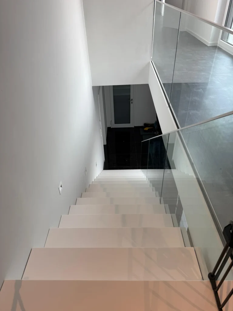 Blick auf eine moderne, weiße Treppe mit Glasgeländer, die zu einer unteren Etage mit schwarzen Fliesen und einer geschlossenen Tür am Ende führt.