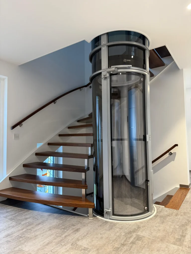 Ein moderner, transparenter zylindrischer Hausaufzug ist neben einer Holztreppe mit Metallgeländer in einem hellen, zeitgenössischen Innenraum installiert.