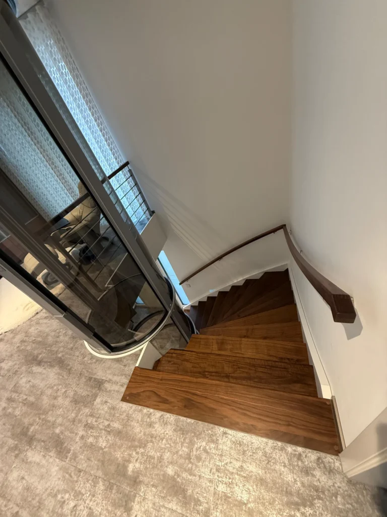 Blick von oben auf eine moderne Wendeltreppe mit Holzstufen und einem eleganten Glasaufzug daneben, umgeben von weißen Wänden und einem Teppichboden.