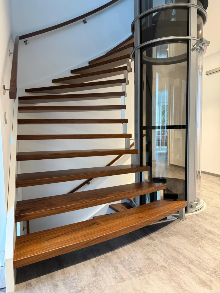 Eine moderne Wendeltreppe mit Holzstufen windet sich um einen zylindrischen Glasaufzug in einem hellen, minimalistischen Innenraum mit hellen Böden und weißen Wänden.