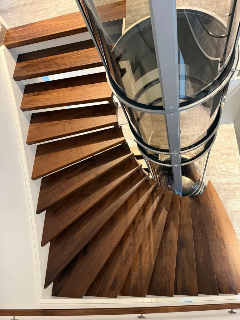 Eine moderne Wendeltreppe mit dunklen Holzstufen schlängelt sich um einen transparenten zylindrischen Aufzug, der von oben auf einem hellen Boden steht.