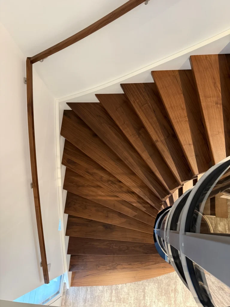 Spindeltreppe mit dunklen Holzstufen und einem glatten, an der weißen Wand befestigten Metallhandlauf, von oben gesehen. Das Treppenhaus windet sich um einen zentralen gläsernen Aufzugsschacht.