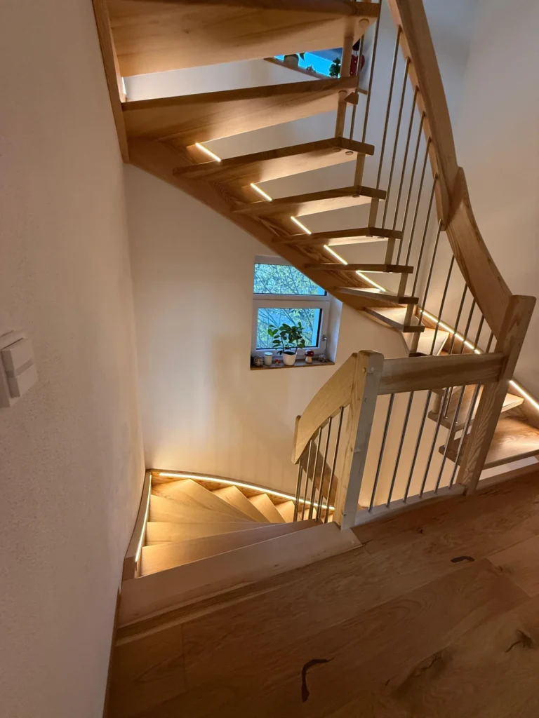 Eine moderne Holztreppe mit eingebauter LED-Beleuchtung entlang der Stufen, die sich um eine Ecke schlängelt. Ein kleines Fenster mit Pflanzen auf der Fensterbank ist an der Wand hinter der Treppe zu sehen.