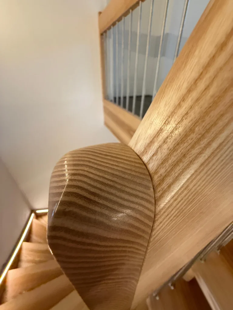 Nahaufnahme eines glatten, geschwungenen hölzernen Handlaufs am oberen Ende einer Treppe mit Metallgeländerstäben und hölzernen Stufen, die die Holzmaserung und die Handwerkskunst zeigen. Der Hintergrund ist sanft unscharf.
