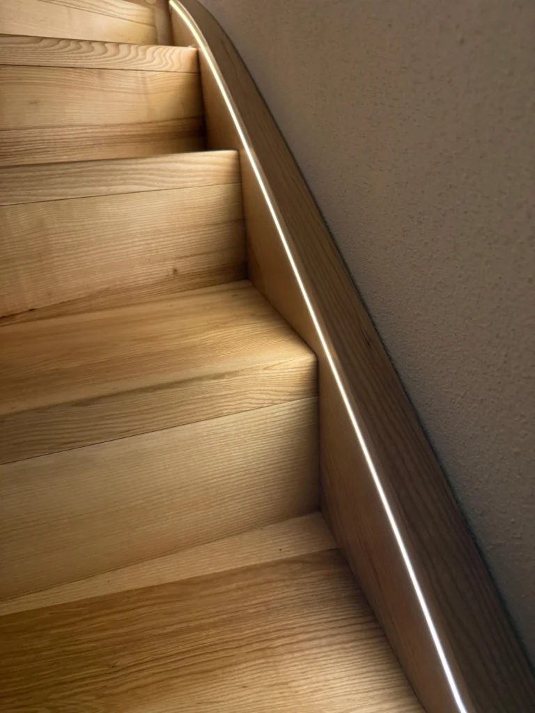 Nahaufnahme einer Holztreppe mit glatter Oberfläche, mit einer geschwungenen Wand auf der rechten Seite und einem dünnen, durchgehenden LED-Lichtstreifen, der entlang der Kante eingebettet ist und ein weiches, modernes Licht erzeugt.