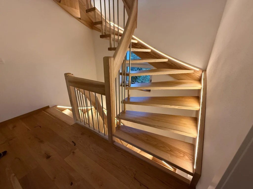 Eine moderne Holztreppe mit offenen Setzstufen und Metallgeländer, beleuchtet von sanften LED-Lichtbändern entlang der Stufen und des Geländers, vor weißen Wänden und Holzböden.