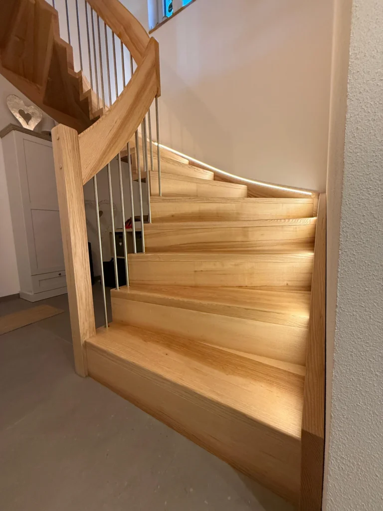 Eine Holztreppe mit eingebauter LED-Beleuchtung entlang der Stufen führt neben einer weißen Wand nach oben. Vertikale Metallgeländerstäbe und Holzhandläufe betonen das moderne, klare Design.