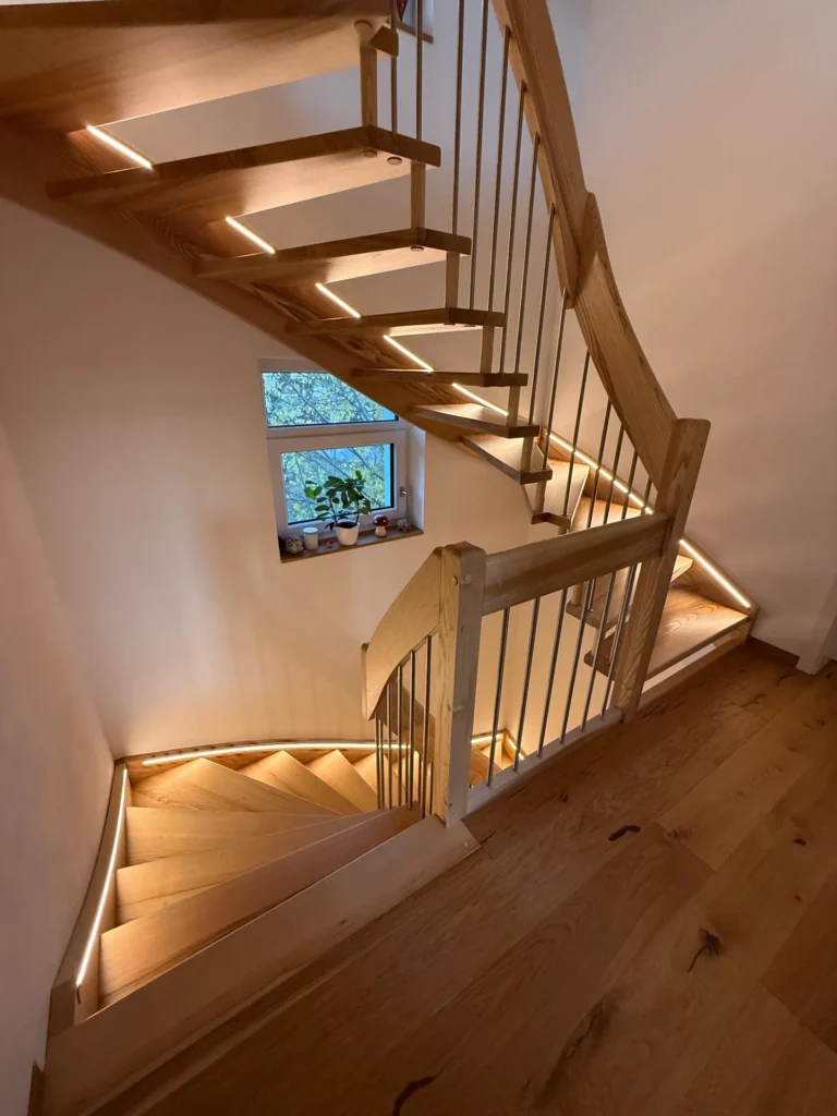 Eine Holztreppe mit LED-Lichtbändern entlang der Stufen schlängelt sich um ein kleines Fenster mit Topfpflanzen auf der Fensterbank. Die Treppe hat ein vertikales Metallgeländer und einen hellen Holzfußboden.