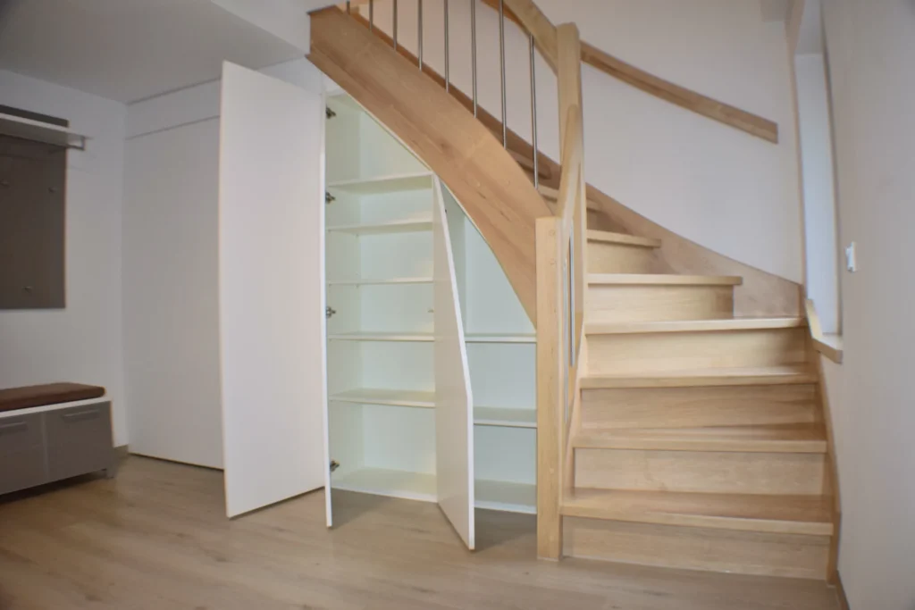 Eine Holztreppe mit geschwungenem Geländer wird neben weißen Schränken mit offenen Türen gezeigt, die den Blick auf Regale freigeben, die in den Raum unter der Treppe als Stauraum eingebaut sind.