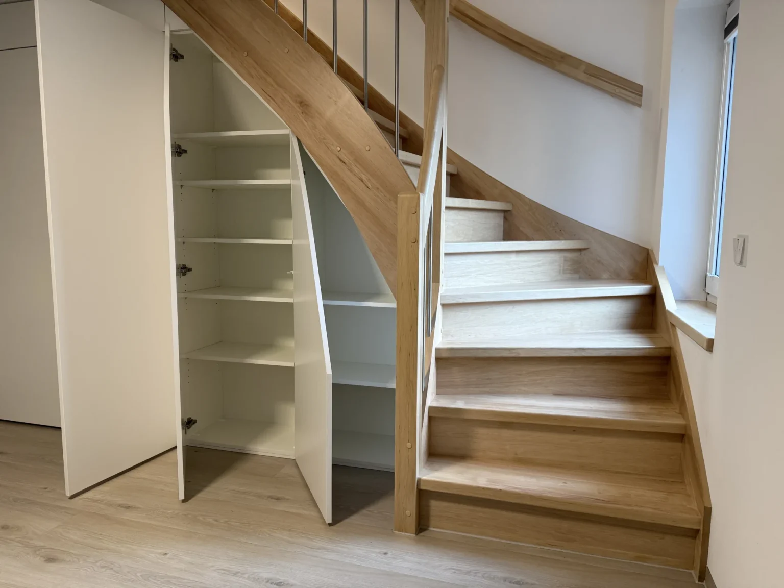 Eine Holztreppe mit weißen Einbauschränken, darunter offene Regale und Türen, in einem modernen, hellen Innenraum mit hellem Holzboden.