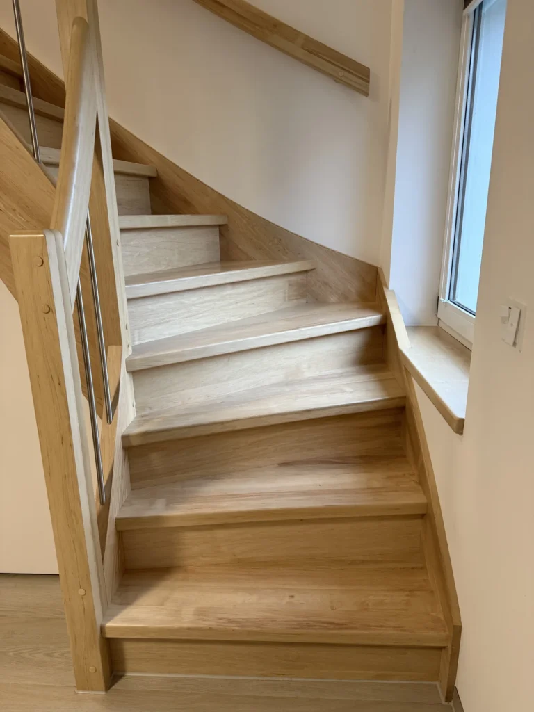 Eine moderne Holztreppe mit hellen Stufen und Handläufen, mit Metallspindeln und einem Fenster auf der rechten Seite, das Tageslicht hereinlässt.