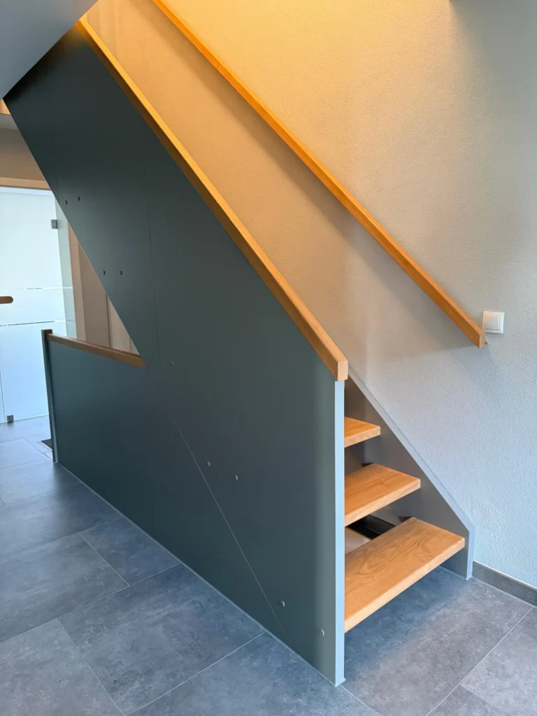 Moderne Innentreppe mit Holzstufen, einer grauen vertikalen Seitenwand und einem hellen Holzhandlauf vor weißen Wänden und grauem Fliesenboden in einem hellen, minimalistischen Raum.